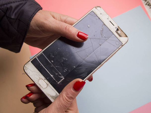 A woman holding a battered broken phone on a neutral multi-colored background. She has no money for repairs and a new manicure. "No money - no honey" concept. Sad modern common situation. - Photo, Image