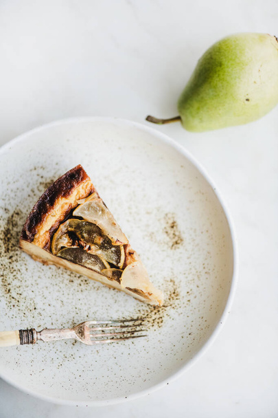 Flat-lay of piece of vegan dairy-free sugar-free pear cheesecake in rustic plate and fresh pear over white marble table background, top view. Modern vegetarian healthy dessert concept - Foto, Imagem