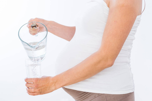 Hamile kadın beyaz arka planda kavanozdan bardağa su döküyor. - Fotoğraf, Görsel