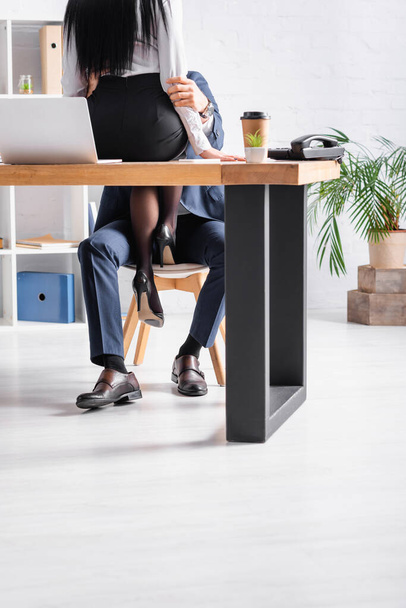 cropped view of businesswoman sitting on desk while seducing colleague in office - Foto, Imagen