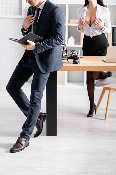 Ausgeschnittene Ansicht einer verführerischen Geschäftsfrau, die eine Bluse aufknöpft, in der Nähe eines Geschäftsmannes, der mit Notizbuch steht - Foto, Bild