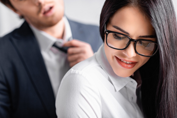 smiling, seductive businesswoman in eyeglasses near colleague adjusting tie on blurred background - Photo, Image