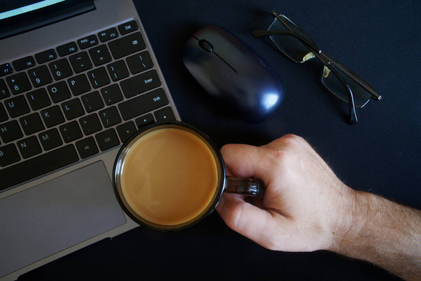 Een man houdt een mok koffie met melk vast terwijl hij achter een moderne laptop of ultraboek zit. Risico van beschadiging en verlies van garantie. Zonder gezicht - Foto, afbeelding