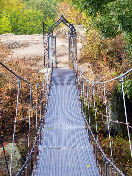 Ανάρτηση μεταλλική γέφυρα πάνω από ένα ποτάμι με υψηλές πέτρινες όχθες. Περπατήστε σε ένα ευχάριστο φθινόπωρο πάρκο σε μια ηλιόλουστη μέρα. Όμορφη σφυρήλατη αψίδα στο τέλος της γέφυρας. - Φωτογραφία, εικόνα
