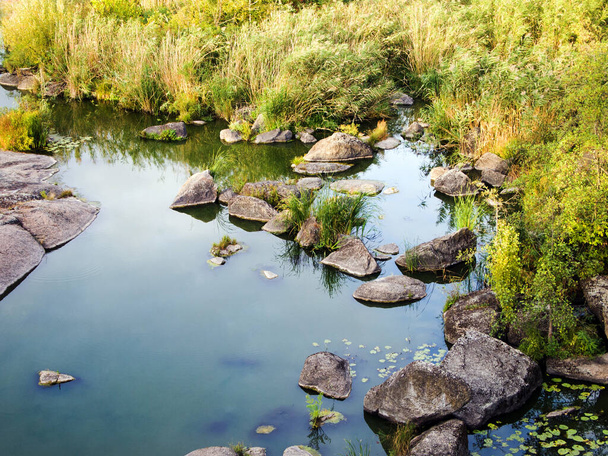 Niesamowita jesienna przyroda: wysoka żółta trawa i duże kamienie w pobliżu brzegu rzeki. Rośliny odbijają się na spokojnej powierzchni wody. Oświetlenie przyciemniające. - Zdjęcie, obraz
