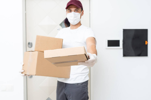 Kurier, Zusteller in medizinischen Latexhandschuhen und Maske liefert während der Coronavirus-Epidemie, COVID-19, sicher Online-Einkäufe in einer weißen Box an die Tür. Zuhause bleiben, sicheres Konzept. - Foto, Bild