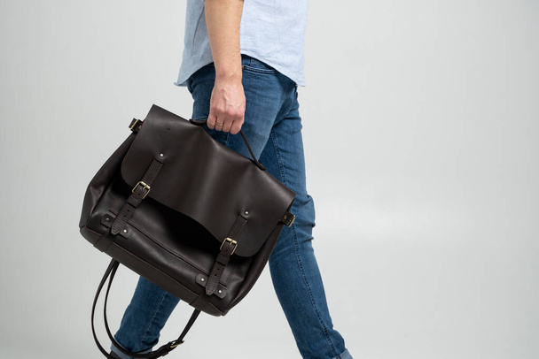 Brown mens shoulder leather bag for a documents and laptop holds by man in a blue shirt and jeans with a white background. Satchel, mens leather handmade briefcase. - Foto, Bild