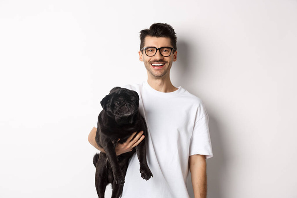 Guapo hipster chico sosteniendo su divertido perro negro pug, sonriendo a la cámara, de pie sobre fondo blanco - Foto, imagen