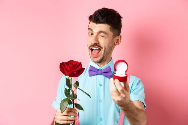 День Святого Валентина. Забавный парень делает предложение, подмигивает и говорит жениться на мне, показывая обручальное кольцо с красной розой, стоя на розовом фоне - Фото, изображение