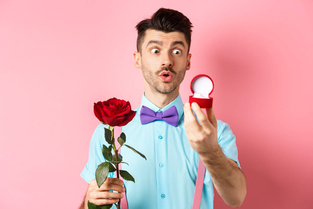バレンタインデー。ロマンチックな面白い男は結婚式の提案をする彼と結婚し、赤いバラを保持し、興奮して婚約指輪を見て、ピンクの背景 - 写真・画像