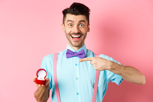 Valentinstag. Fröhlicher junger Mann macht Heiratsantrag, zeigt mit dem Finger auf Verlobungsring und lächelt aufgeregt, steht auf romantischem rosa Hintergrund - Foto, Bild