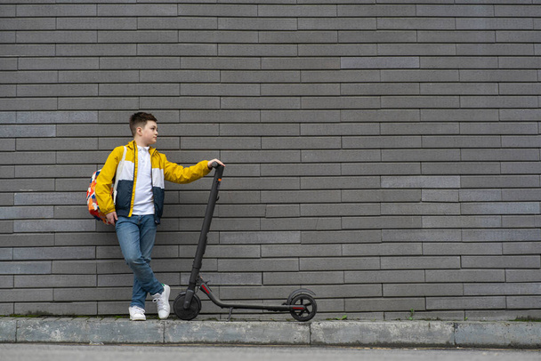 Tiener met rugzak en elektrische scooter op bakstenen muur achtergrond. Modern schooljongensconcept - Foto, afbeelding