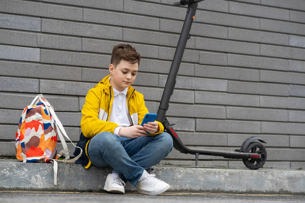 Tiener met mobiele telefoon naast zijn elektrische scooter en rugzak. Moderne jonge jongen chatten op smartphone - Foto, afbeelding
