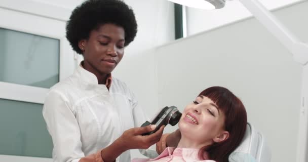 El médico es dermatólogo afroamericano examina una piel. Médico profesional El dermatólogo investiga una marca de nacimiento en el teléfono conectado a la herramienta especial de dermatoscopia. Examen de la cara - Imágenes, Vídeo