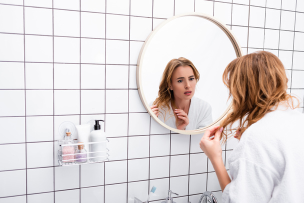 беспокойная женщина в халате регулирует волосы, глядя на зеркало в ванной комнате  - Фото, изображение