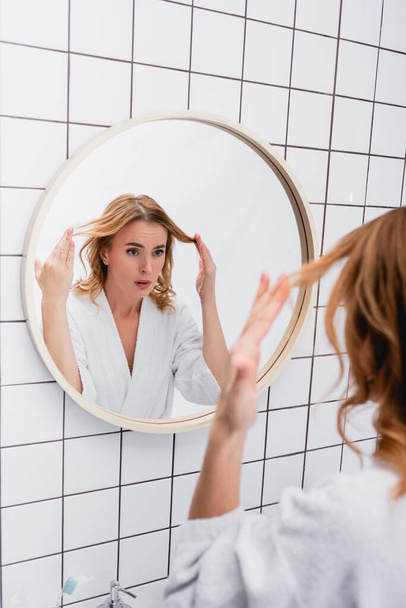 displeased woman in bathrobe adjusting hair while looking at mirror in bathroom  - Foto, Imagen