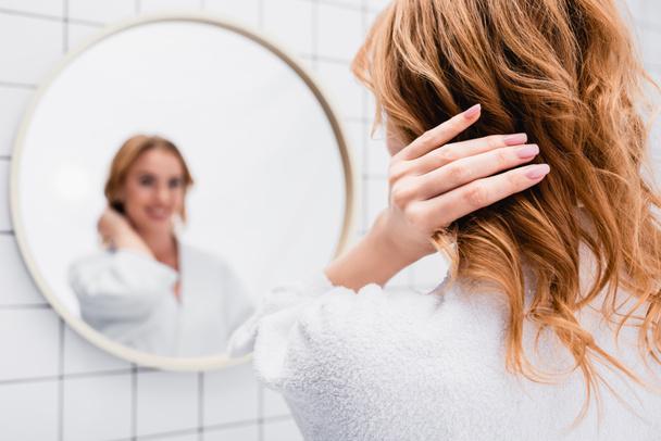 kobieta w szlafroku regulacja włosów w pobliżu lustra na rozmytym tle - Zdjęcie, obraz