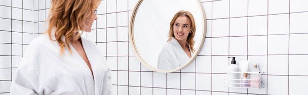 vidám nő fürdőköpenyben mosolyog, miközben nézi a tükröt a fürdőszobában, banner - Fotó, kép