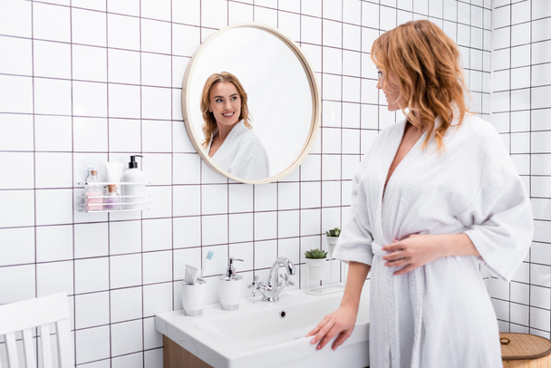joyful woman in bathrobe smiling while looking at mirror in bathroom - Foto, afbeelding