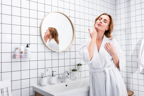 mujer con los ojos cerrados aplicando crema facial en el baño - Foto, imagen
