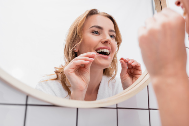 reflejo de la mujer feliz mirando al espejo y sosteniendo hilo dental - Foto, Imagen