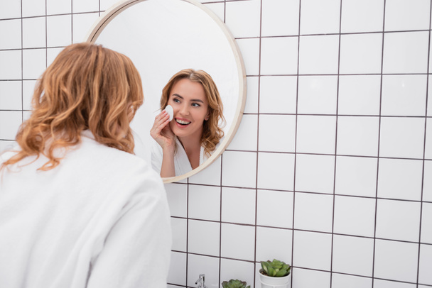 щаслива жінка наносить тонер з ватним майданчиком на обличчя і дивиться на дзеркало у ванній
  - Фото, зображення