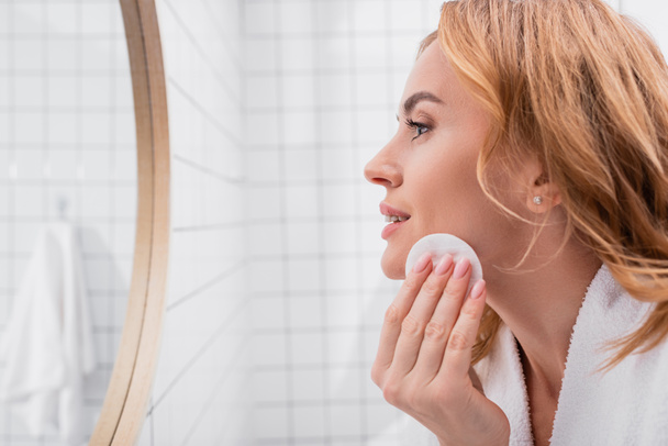 vista lateral de la mujer aplicando tóner con almohadilla de algodón en la cara y mirando el espejo  - Foto, Imagen