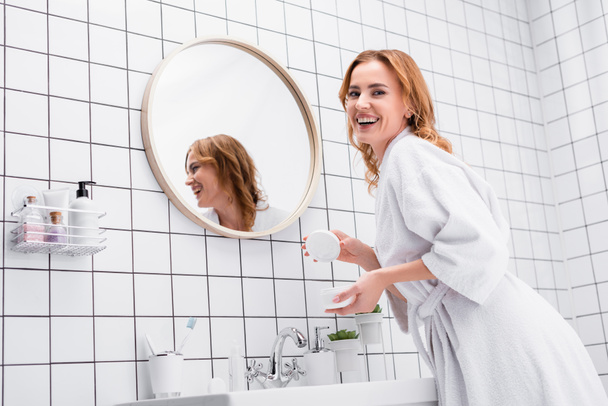 mujer alegre sosteniendo tarro con crema facial y mirando a la cámara en el baño  - Foto, Imagen