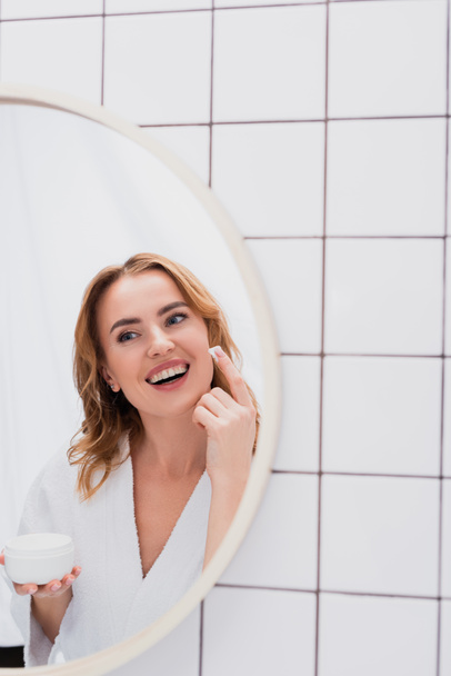 mujer alegre aplicación de crema facial mientras mira el espejo  - Foto, Imagen
