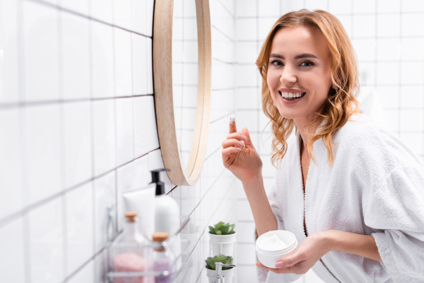 sonriente mujer sosteniendo frasco con crema facial cerca del espejo cerca de botellas en primer plano borrosa  - Foto, Imagen