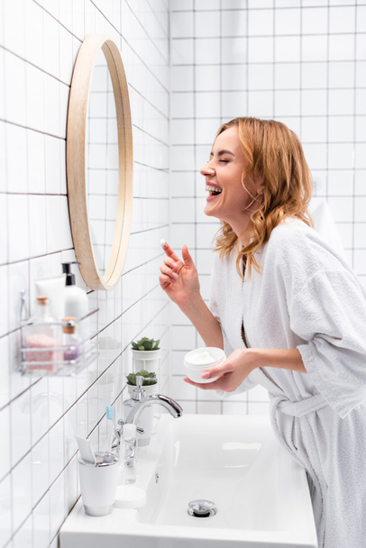 улыбающаяся женщина держит банку с кремом для лица и смеется возле зеркала и бутылки на размытом переднем плане  - Фото, изображение