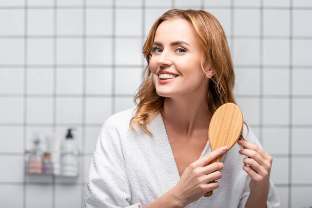 ευτυχισμένη γυναίκα σε λευκό μπουρνούζι βούρτσισμα των μαλλιών στο μπάνιο - Φωτογραφία, εικόνα