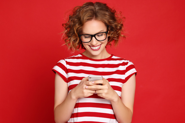 Verrukte jonge vrouw in gestreept t-shirt en trendy bril lezen onverwachte boodschap op smartphone met glimlach tegen rode achtergrond - Foto, afbeelding