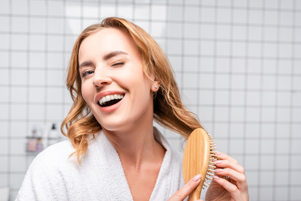 iloinen nainen hymyilee ja iskee silmää harjaamalla hiuksia kylpyhuoneessa - Valokuva, kuva
