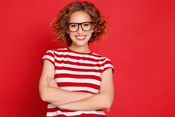 Selbstbewusste junge Frau in gestreiftem T-Shirt und Brille, die die Arme verschränkt und vor rotem Hintergrund für die Kamera lächelt - Foto, Bild