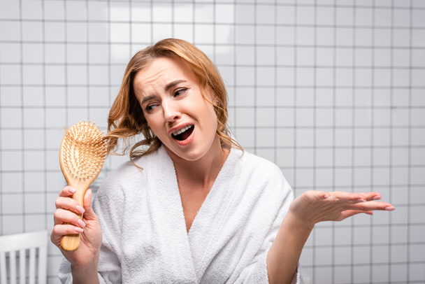 δυσαρεστημένη γυναίκα με μπουρνούζι βουρτσίζει τα μαλλιά στο μπάνιο - Φωτογραφία, εικόνα