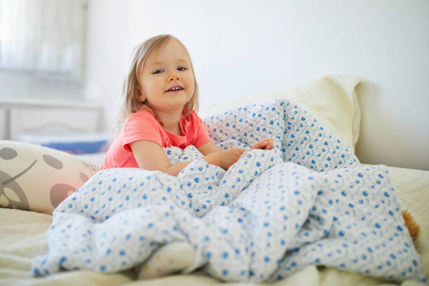 Happy toddler girl in striped red and white pajamas sitting on bed right after awaking. Day naps for small kids - Фото, зображення