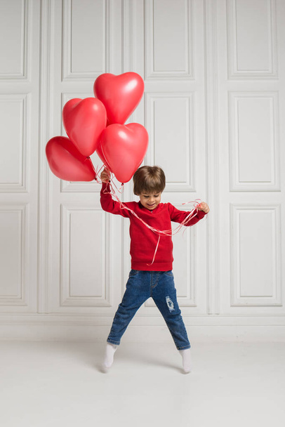 un petit garçon en jean et un pull saute avec des ballons rouges sur un fond blanc avec de l'espace pour le texte - Photo, image