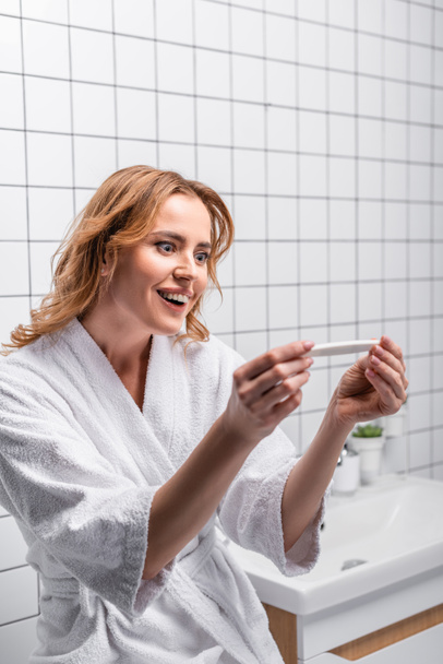 glückliche Frau im weißen Bademantel hält Schwangerschaftstest im Badezimmer - Foto, Bild