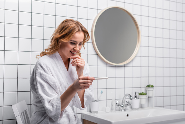 örömteli nő fehér fürdőköpenyben nézi a terhességi tesztet a fürdőszobában - Fotó, kép