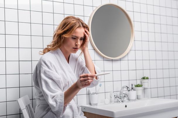 worried woman in white bathrobe holding pregnancy test in bathroom - Foto, Bild