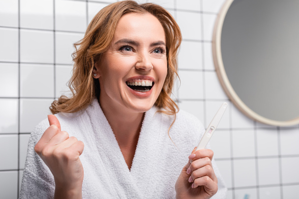 aufgeregte Frau im weißen Bademantel hält Schwangerschaftstest im Badezimmer - Foto, Bild