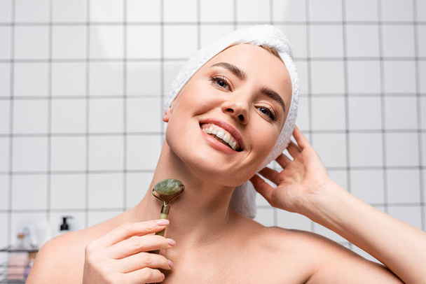 fröhliche Frau mit Handtuch auf dem Kopf mit Jadewalze im Badezimmer - Foto, Bild