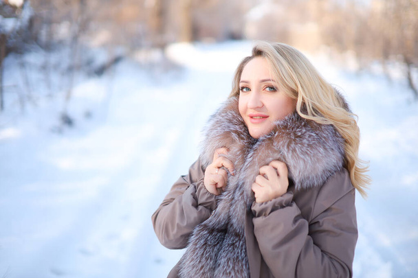 una rubia en una chaqueta con un cuello de piel se para de lado en un parque de invierno - Foto, imagen