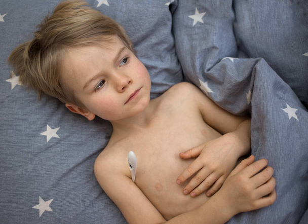 Αρρωστημένο παιδί, ξαπλώνει σε ένα μαξιλάρι στο κρεβάτι με θερμόμετρο. το παιδί μετρά τη θερμοκρασία του σώματος στο κρεβάτι, δεν αισθάνεται καλά - Φωτογραφία, εικόνα
