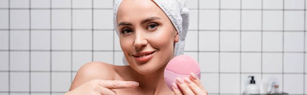 cheerful woman in towel on head pointing with finger at cleansing silicone brush in bathroom - Photo, Image