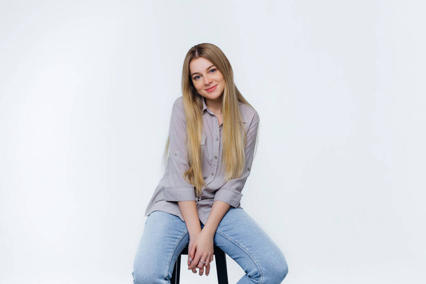 krásná roztomilá dívka usměvavá sedí na židli ve studiu, oblečená v džínách a blůze s blond dlouhými vlasy. - Fotografie, Obrázek
