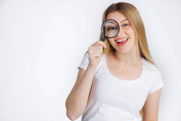 Funny joyful girl with beautiful long blond hair in a white t-shirt looking through a magnifying glass - Photo, Image