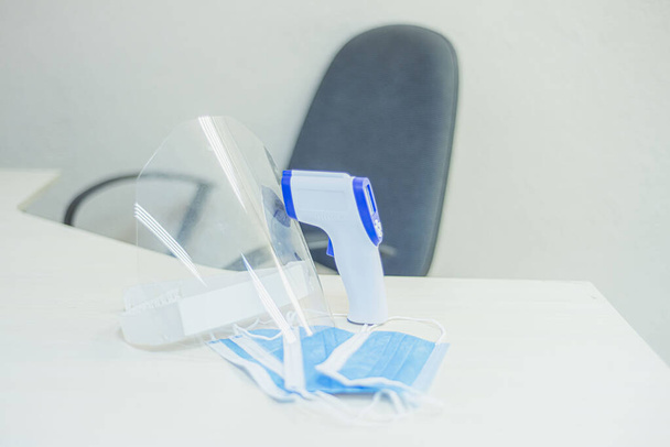 защитная маска и термометр на столе, измерить температуру в офисе, карантин, пустой стул, удаленная работа - Фото, изображение