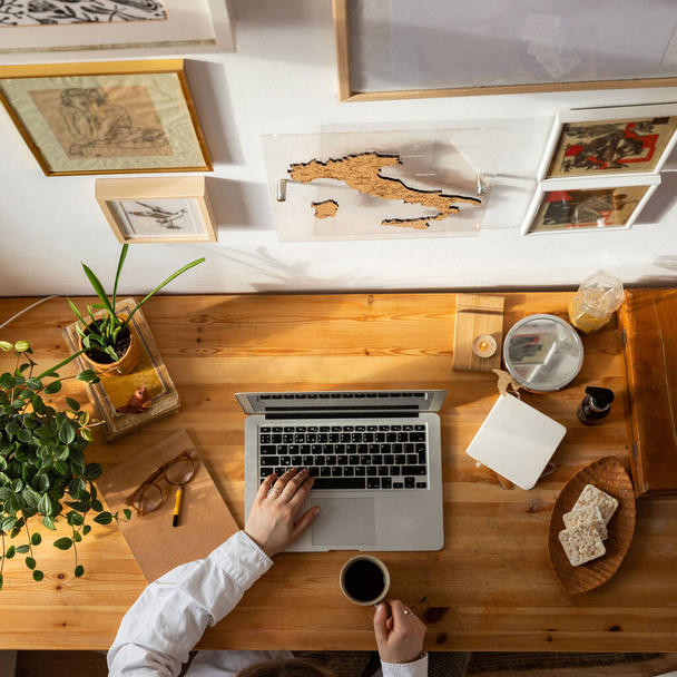 Drewniany widok z góry, kobieta projektant ręce za pomocą laptopa i trzymając filiżankę kawy. Miejsce pracy freelancera - rośliny domowe, ciasteczka, obraz na ścianie, szkicownik, rocznik książki. Praca w domu. - Zdjęcie, obraz